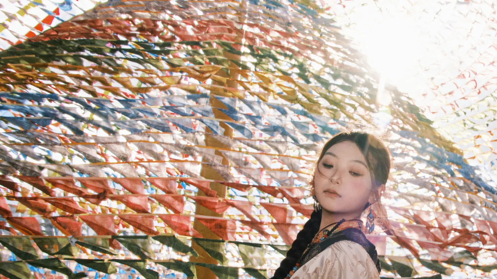 女子站在经幡下仰头，逆光穿过彩旗