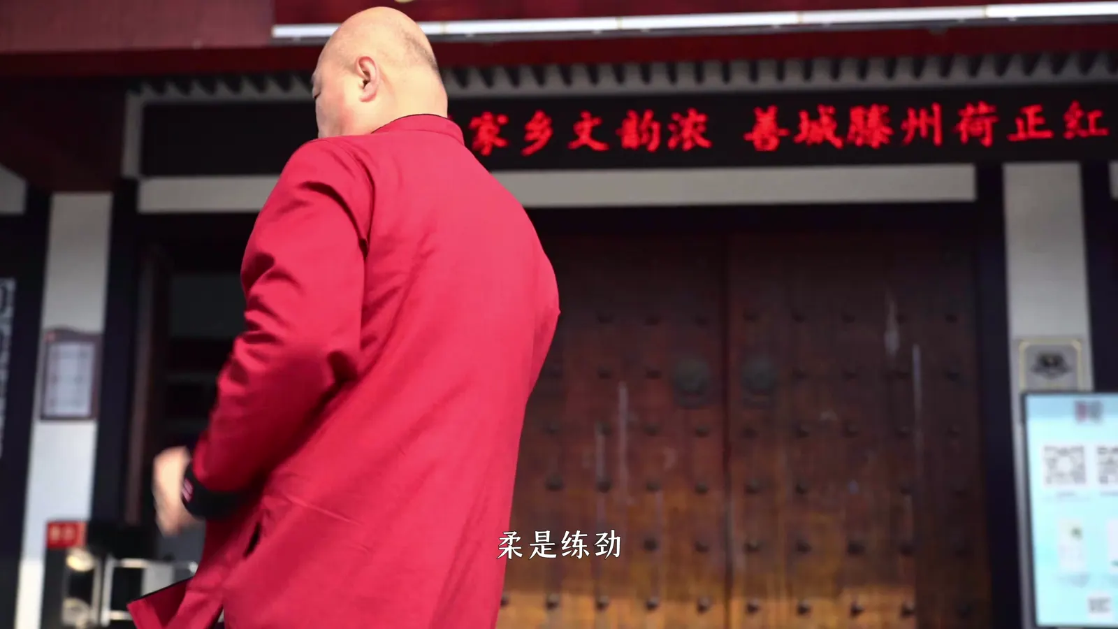 身穿红色练功服的老者在老宅门前转身，屋檐下悬着红底横匾